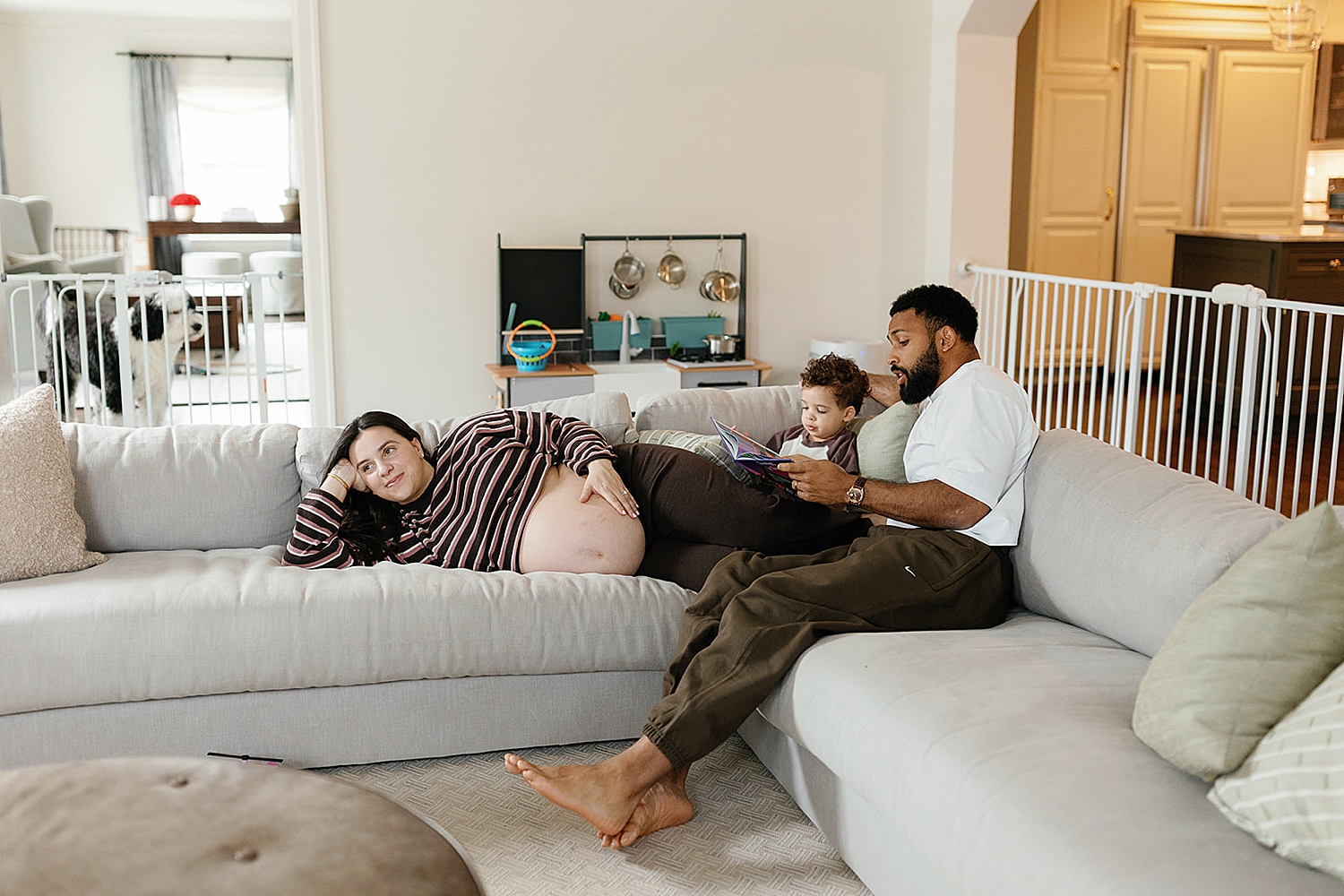 pregnant woman lies across couch holding her bump with her fam nearby by Rachel Andrade Photography