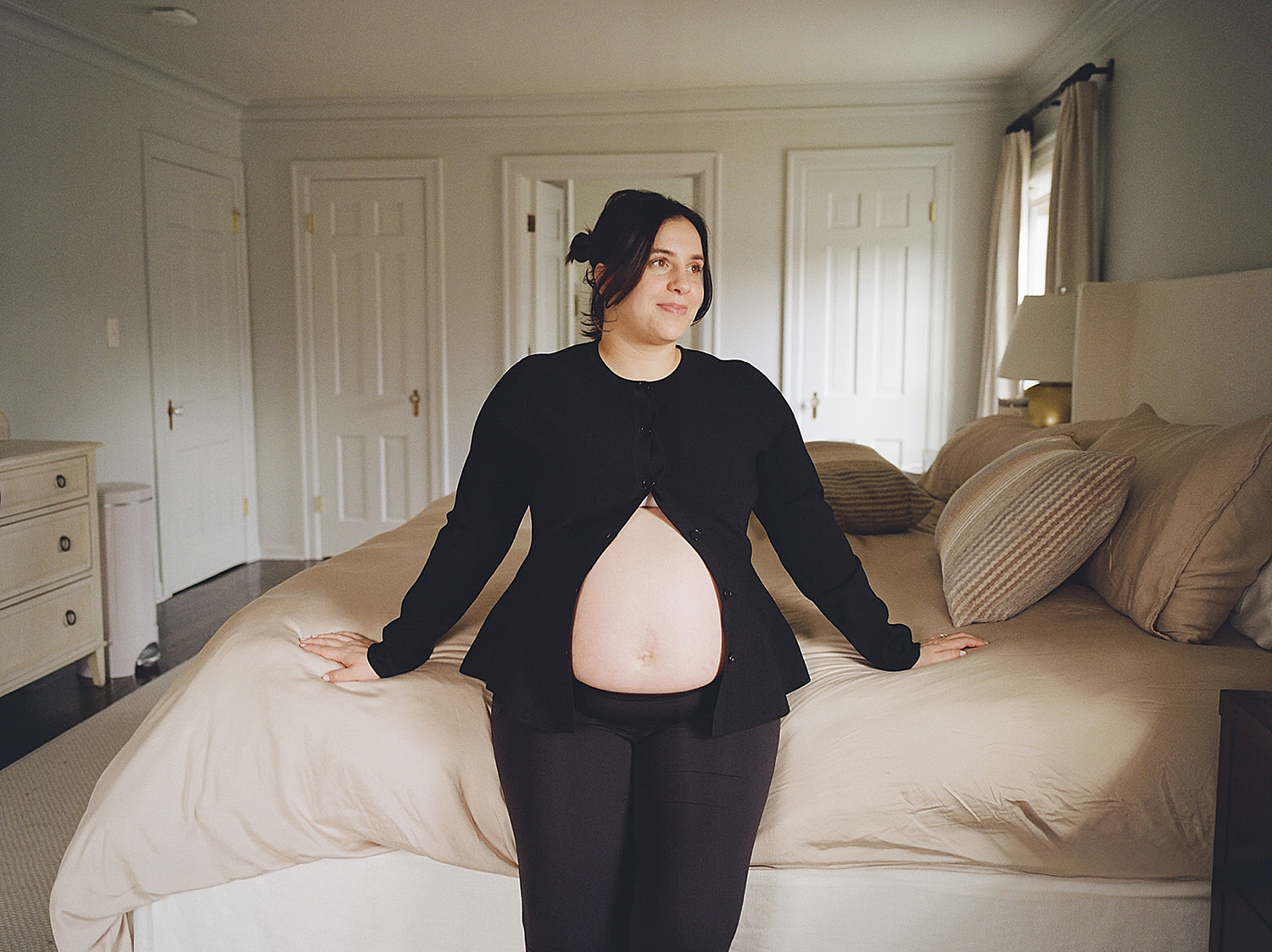 woman with baby bump leans against their bed by Rachel Andrade Photography